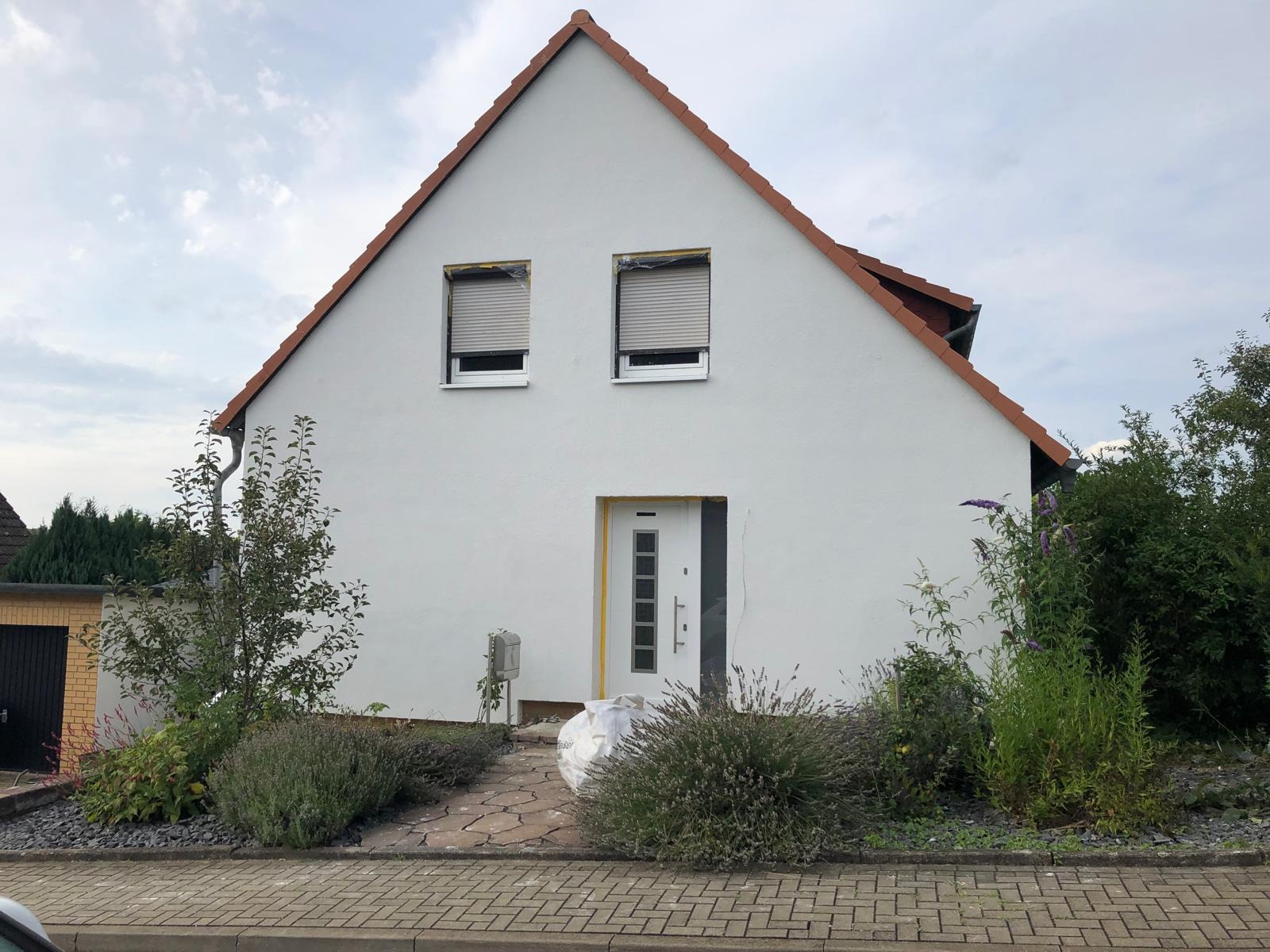 Biały dom jednorodzinny z czerwonym dachem, okna zabezpieczone folią malarską, widoczna barierka ochronna wokół drzwi wejściowych i workiem z odpadami budowlanymi przed wejściem, sugerujące prace...