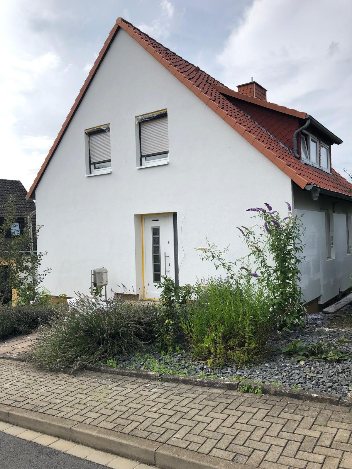 Dom jednorodzinny z nowo pomalowaną białą elewacją, okna zabezpieczone folią, widoczne rośliny w ogrodzie i chodnik z kostki brukowej.