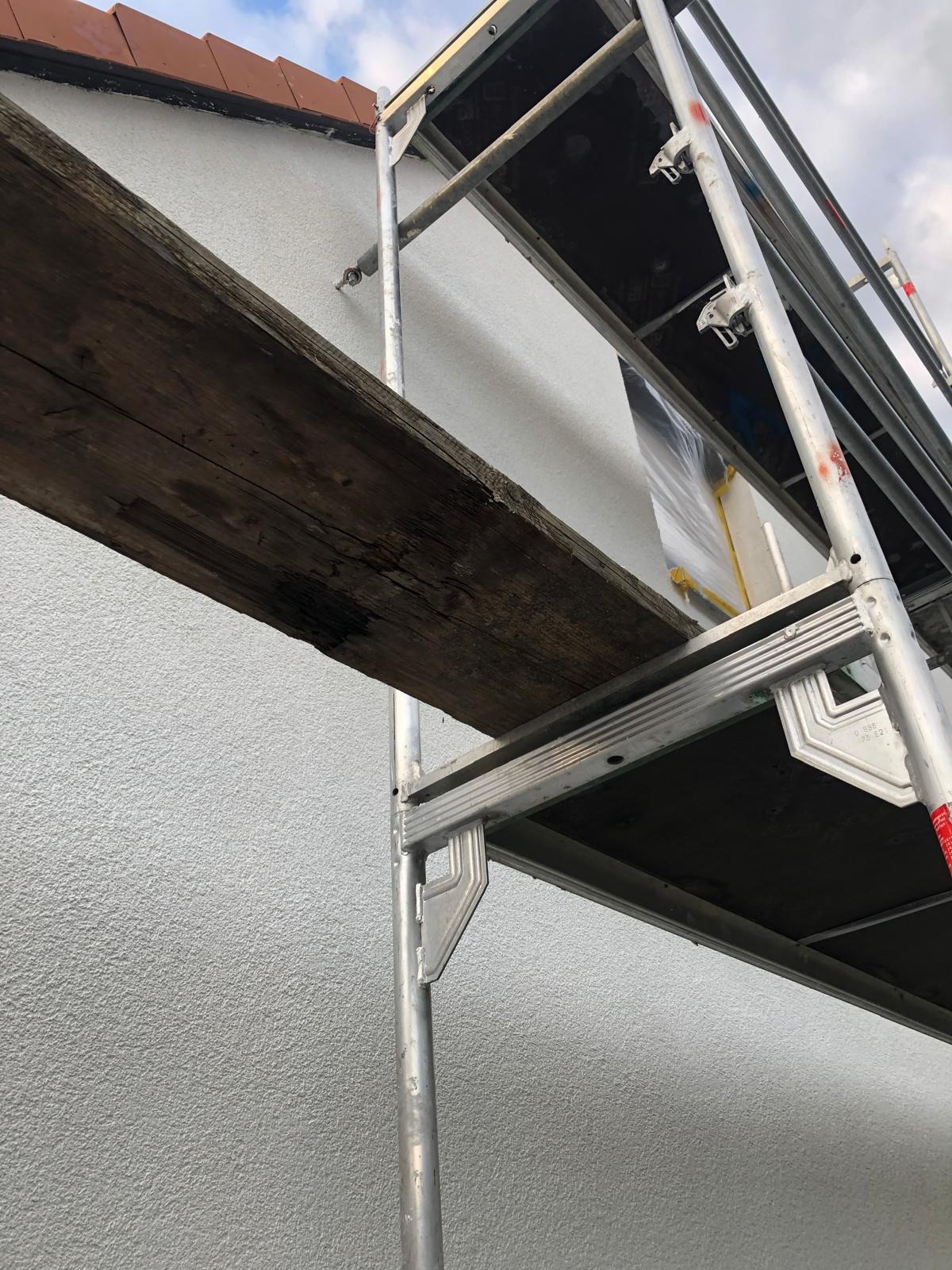 Szare rusztowanie z drewnianą deską na tle świeżo pomalowanej, jasnoszarej elewacji z widoczną fakturą. Częściowo widoczny dach z czerwoną dachówką.