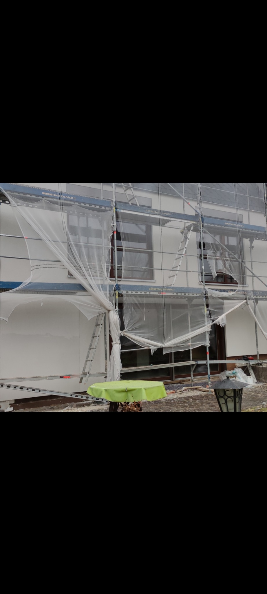 Elewacja domu w trakcie prac, osłonięta siatką na rusztowaniu, z widoczną drabiną i zielonym stolikiem na pierwszym planie.