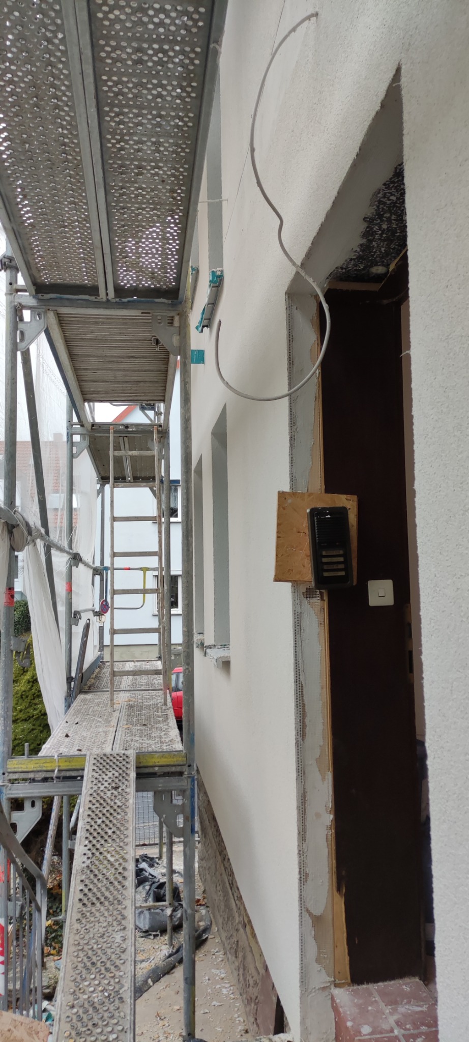 Elewacja budynku w trakcie prac: świeżo otynkowana ściana z widocznym ościeżem drzwi wejściowych, przewody elektryczne wystające ze ściany, rusztowanie z metalową kratą w tle.
