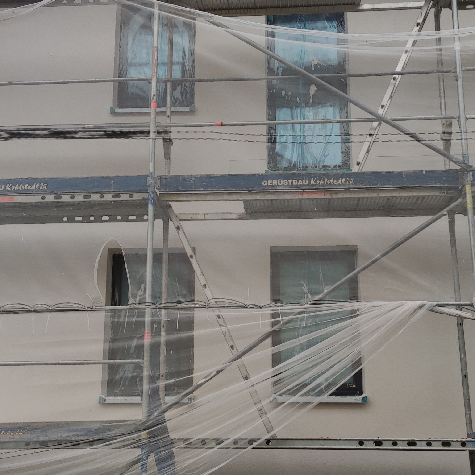 Elewacja budynku w trakcie prac, osłonięta siatką i rusztowaniem, okna zabezpieczone folią.