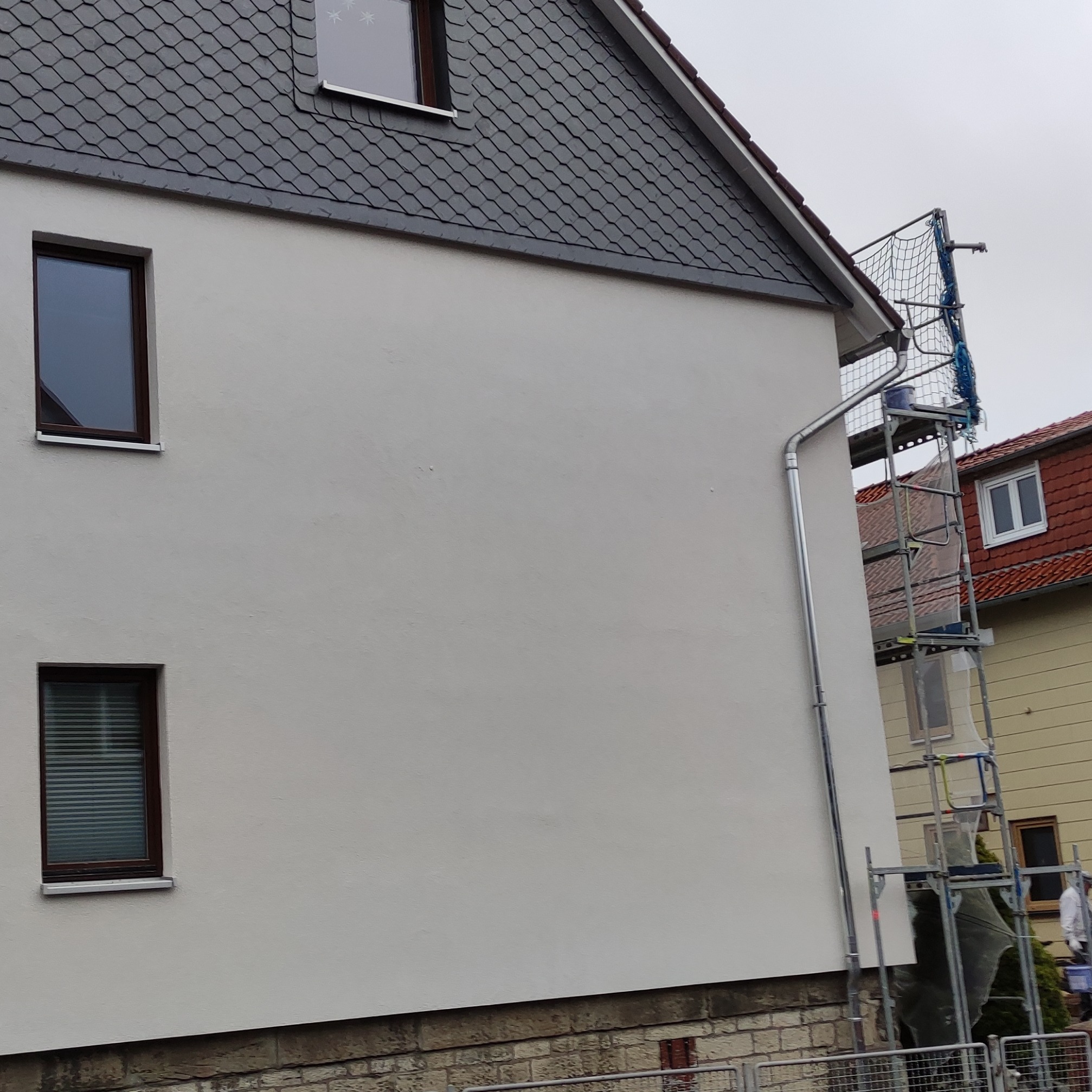 Świeżo pomalowana ściana budynku z szarym dachem i oknami w brązowych ramach, widoczne rusztowanie z niebieską siatką zabezpieczającą i rynna.