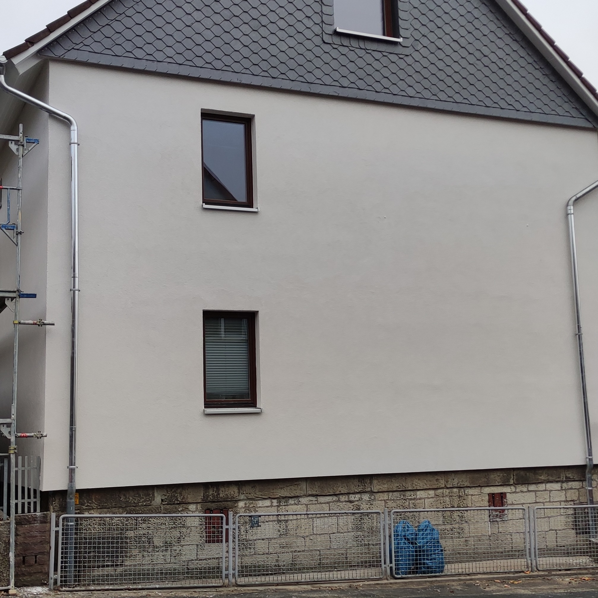 Świeżo pomalowana elewacja domu jednorodzinnego w odcieniach bieli, z trzema oknami o brązowych ramach i szarym dachem z dachówki łuskowej. Widoczne rynny i fragment rusztowania po lewej stronie.
