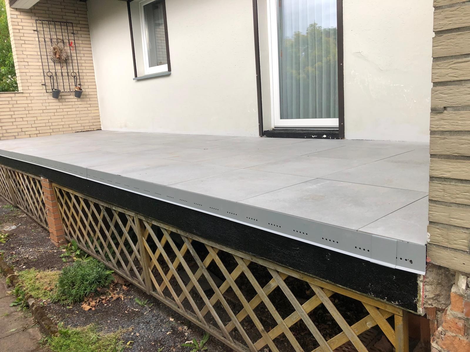 Wykończony taras z szarych płytek, obudowany drewnianą kratownicą i szarą listwą z otworami wentylacyjnymi, widok z poziomu gruntu.