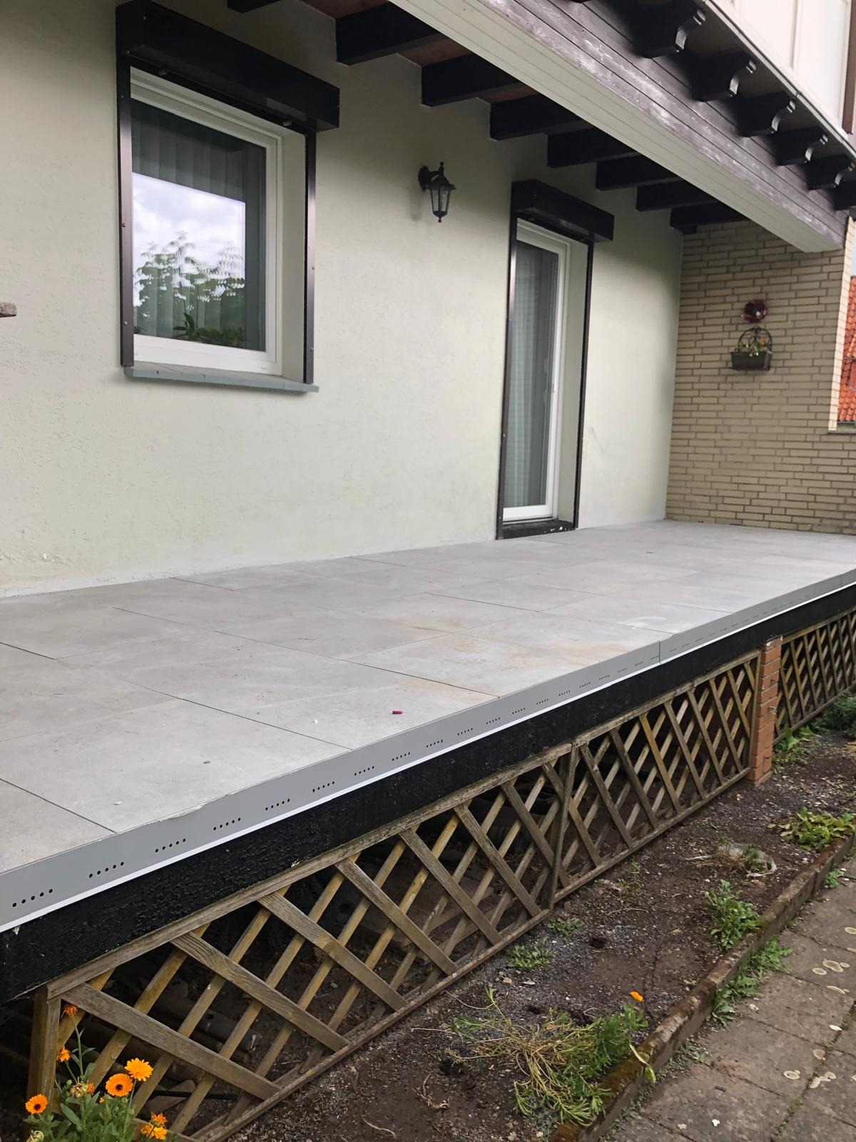 Wyłożony szarymi płytkami taras przed domem z drewnianą kratką maskującą podkonstrukcję, widoczne okna z roletami zewnętrznymi i lampa na elewacji.