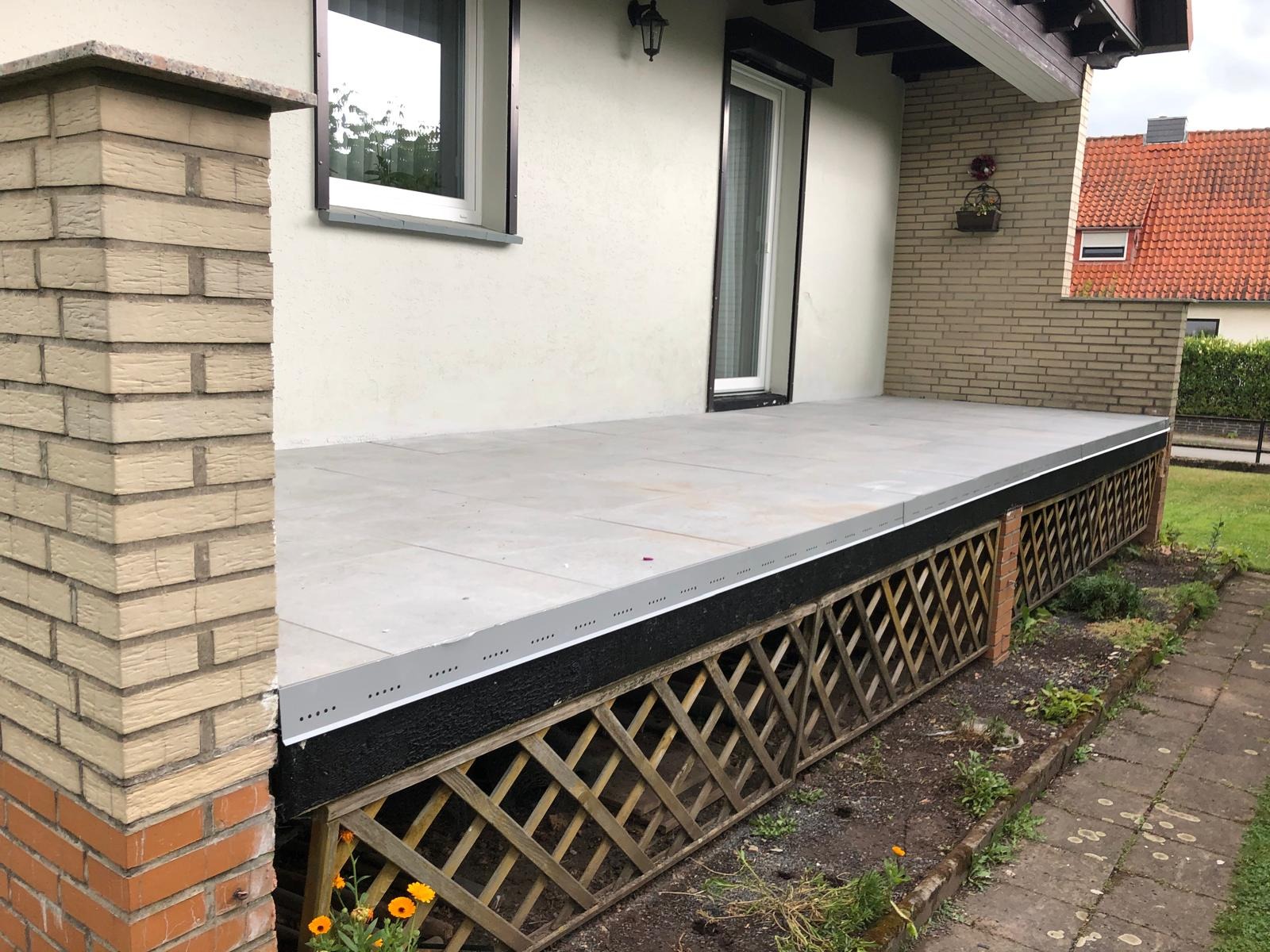 Zadaszony taras wyłożony jasnoszarą płytką, z drewnianą kratownicą maskującą przestrzeń pod spodem, widok z boku, w tle ściana budynku z oknami.