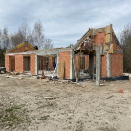 BUDCZEST Sebastian Słaboń - Budowa domu jednorodzinnego w trakcie wznoszenia ścian z czerwonej cegły, widoczne szalunki i zbrojenia stropu, na zewnątrz teren budowy z piaskiem i narzędziami.