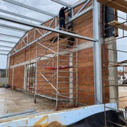 Budowa hali: murowane ściany z pustaków ceramicznych, stalowa konstrukcja dachu, fundamenty z izolacją, pracownik na rusztowaniu.