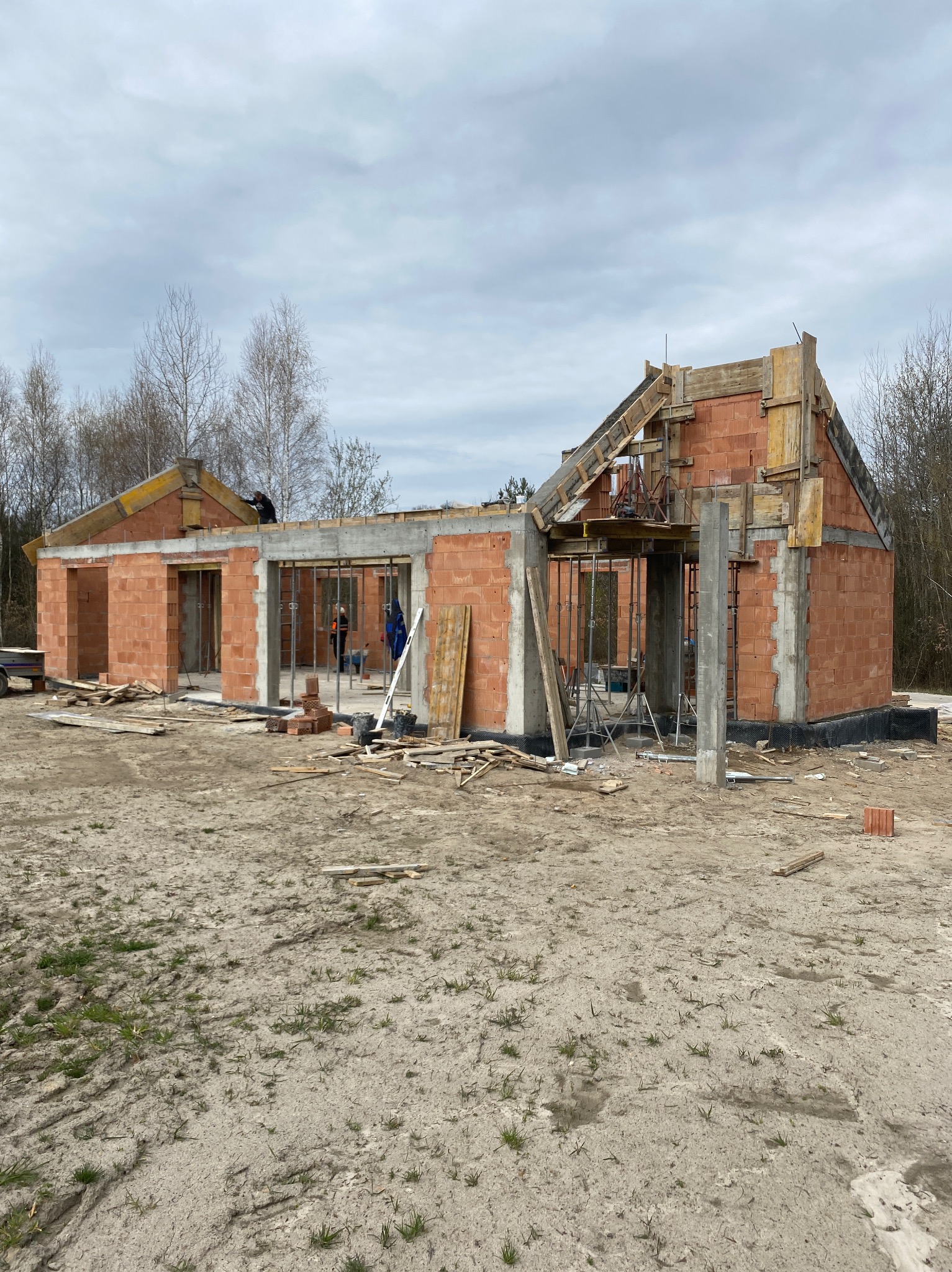 Budowa domu jednorodzinnego w trakcie wznoszenia ścian z czerwonej cegły, widoczne szalunki i zbrojenia stropu, na zewnątrz teren budowy z piaskiem i narzędziami.