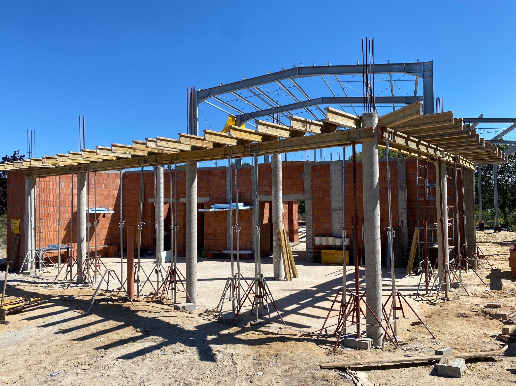 Budowa budynku z cegły: wznoszone ściany z otworami okiennymi, betonowe kolumny podtrzymujące drewniane szalunki stropu, stalowa konstrukcja dachu w tle, widoczne podpory budowlane.