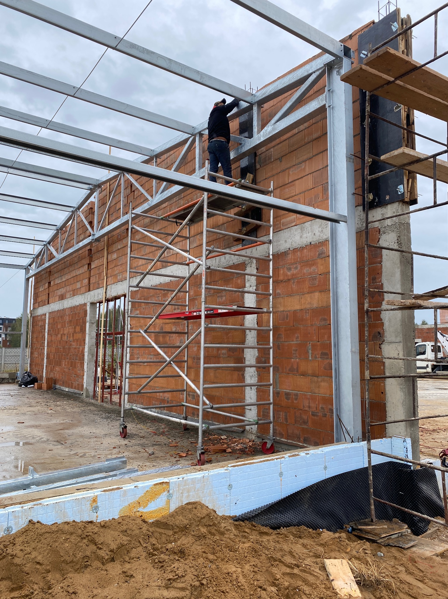 Budowa hali: murowane ściany z pustaków ceramicznych, stalowa konstrukcja dachu, fundamenty z izolacją, pracownik na rusztowaniu.