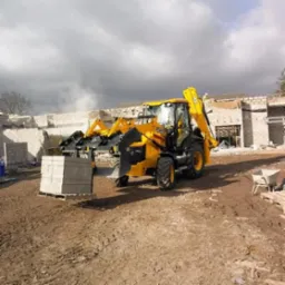 Żółta koparko-ładowarka JCB transportuje paletę z bloczkami betonowymi na placu budowy, w tle wznoszone ściany z pustaków, pochmurne niebo.