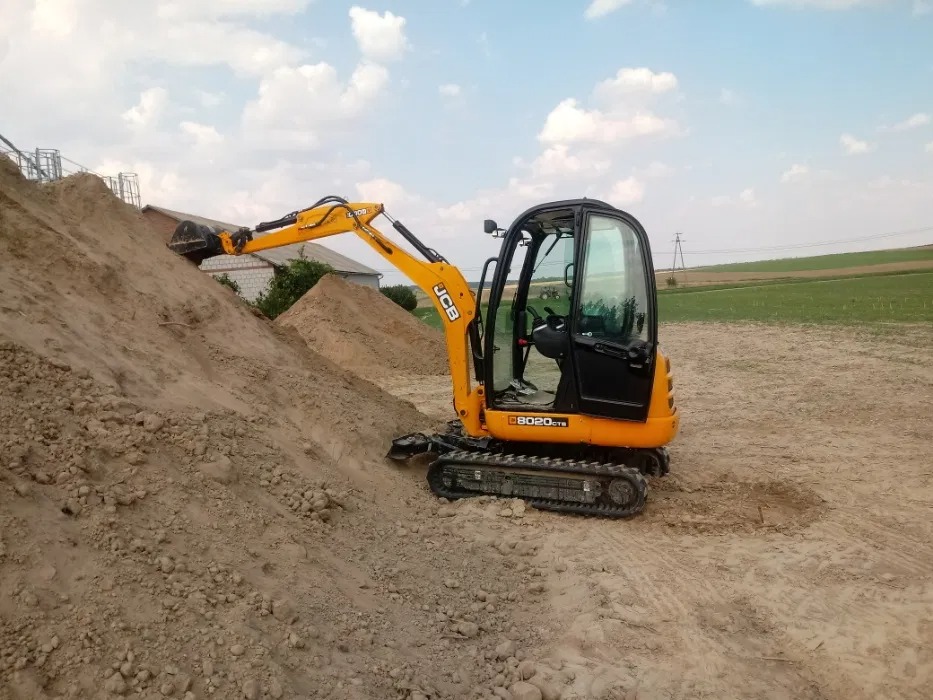 Żółto-czarna mini koparka JCB 8020 CTS nabiera ziemię z dużej pryzmy na placu budowy, w tle budynki i pola uprawne.