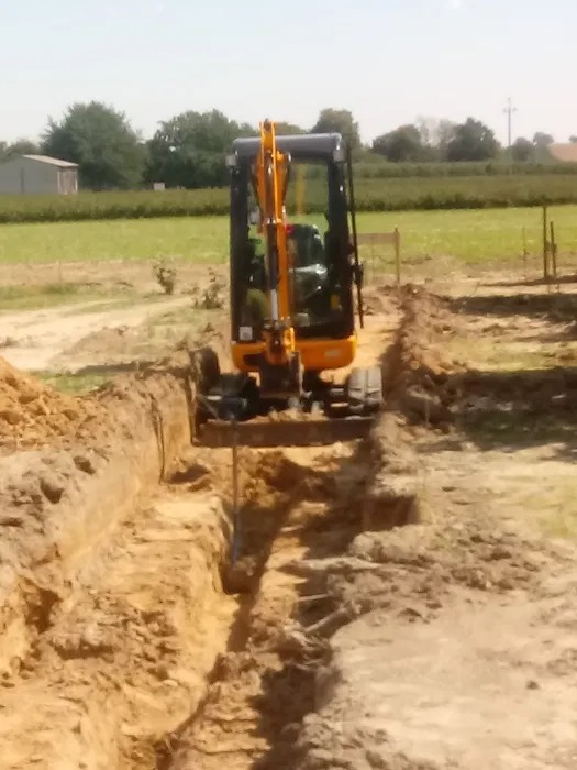 Żółta mini koparka w Świdniku wykonuje wąski wykop w ziemi, widok z przodu na tle pola i zabudowań gospodarczych.