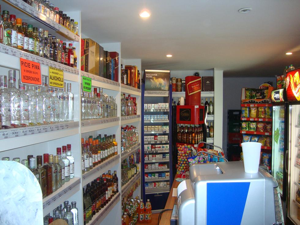 Wnętrze sklepu z alkoholem w Tychach, półki wypełnione różnorodnymi butelkami wódek, win, piw i innych napojów alkoholowych, widoczne również regały z papierosami i przekąskami.
