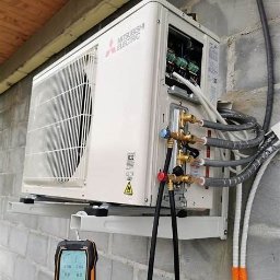 CWK - KLIMA KASPER CIESZKOWSKI - Biała jednostka zewnętrzna klimatyzacji Mitsubishi Electric zamontowana na ścianie z bloczków betonowych, widoczne podłączenia rur i manometry pomiarowe.