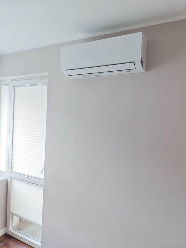 Biały klimatyzator ścienny zamontowany w jasnym pokoju obok okna z plisowanymi roletami.