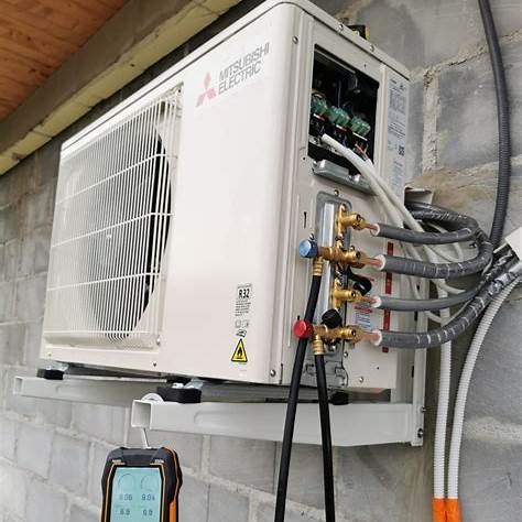 Biała jednostka zewnętrzna klimatyzacji Mitsubishi Electric zamontowana na ścianie z bloczków betonowych, widoczne podłączenia rur i manometry pomiarowe.