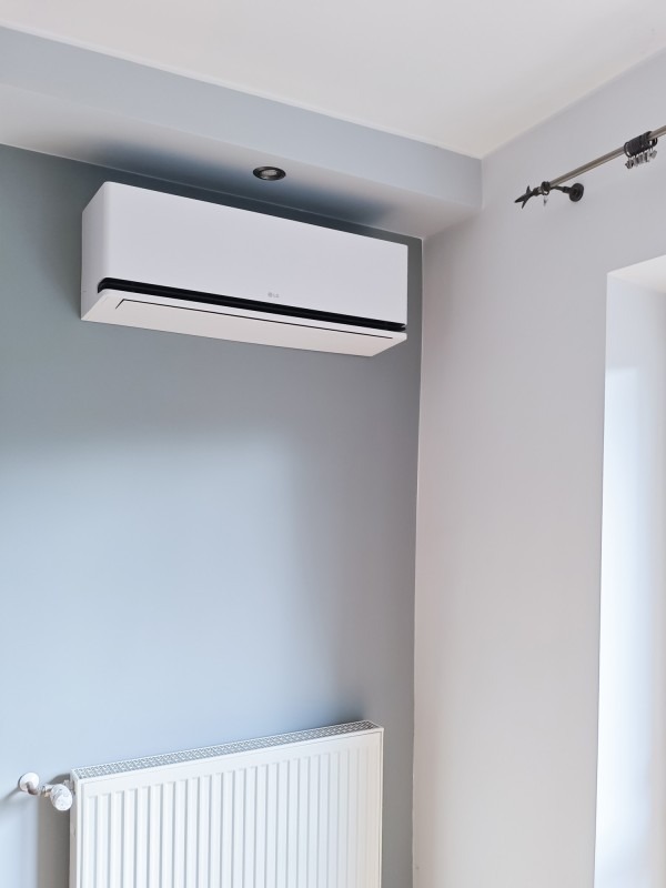 Biały klimatyzator LG zamontowany na szarej ścianie nad białym grzejnikiem w jasnym pomieszczeniu z elementami dekoracyjnymi.