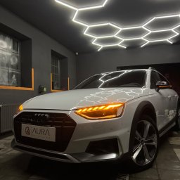 AURA DETAILING - Białe Audi A4 Allroad po detailingu, z włączonymi światłami, widoczne logo 'Aura Detailing', oświetlenie heksagonalne na suficie, czyste wnętrze garażu.