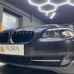 AURA DETAILING - Szary samochód marki BMW po detailingu, widok z przodu, z logo firmy Aura Detailing. W tle widoczne oświetlenie warsztatowe.
