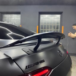 AURA DETAILING - Matowy, czarny Mercedes-AMG z wyraźnie widocznym spojlerem i tylnymi światłami. W tle osoba robiąca zdjęcie telefonem, co sugeruje etap dokumentacji po usłudze.