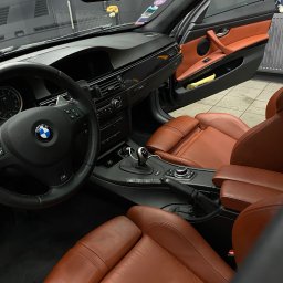 AURA DETAILING - Wnętrze BMW E92 z brązową skórzaną tapicerką, widok na fotele, kierownicę i deskę rozdzielczą. Otwarta boczna szyba. Detailing wnętrza.
