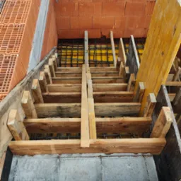 Wykop z deskowaniem schodów, zbrojenie i częściowo wymurowane ściany z cegły dziurawki. Widok z góry na placu budowy w słoneczny dzień.