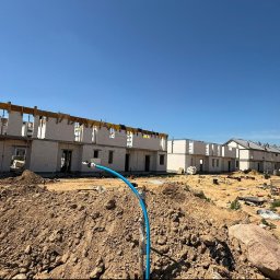 VESTRA HOME SPÓŁKA Z OGRANICZONĄ ODPOWIEDZIALNOŚCIĄ - Wznoszone domy szeregowe z jasnych bloczków na placu budowy, z widocznymi drewnianymi szalunkami stropowymi. Na pierwszym planie hałdy ziemi i niebieska rura instalacyjna pod bezchmurnym niebem.