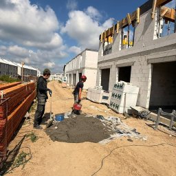 VESTRA HOME SPÓŁKA Z OGRANICZONĄ ODPOWIEDZIALNOŚCIĄ - Dwóch pracowników budowlanych miesza zaprawę na ziemi, obok wznoszonych domów szeregowych z białych bloczków H+H, z widocznymi drewnianymi elementami konstrukcyjnymi. Słoneczny dzień.