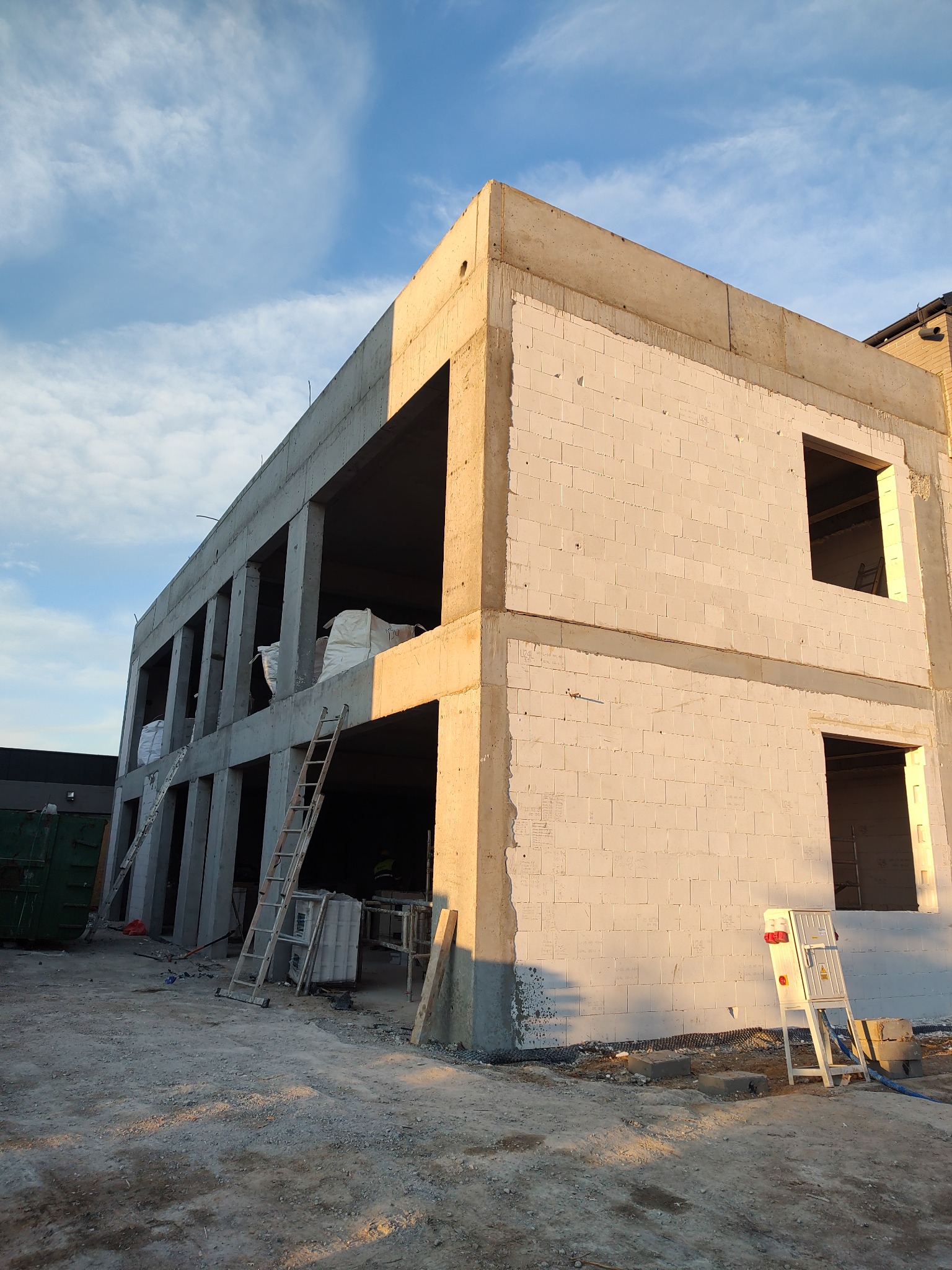 Dwukondygnacyjny budynek w stanie surowym otwartym z betonu i bloczków, widoczne worki z materiałami na piętrze, oświetlony popołudniowym słońcem.