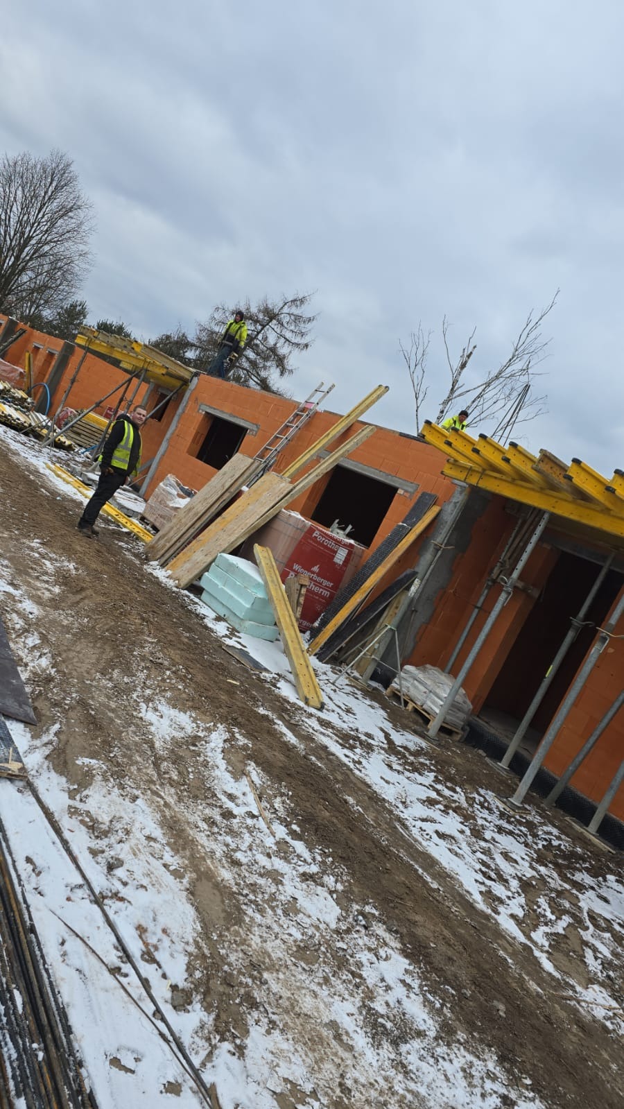 Budowa domu w trakcie prac murarskich, widoczne ściany z bloczków ceramicznych Porotherm, drewniane szalunki, pracownicy w kamizelkach odblaskowych, teren budowy z resztkami śniegu.