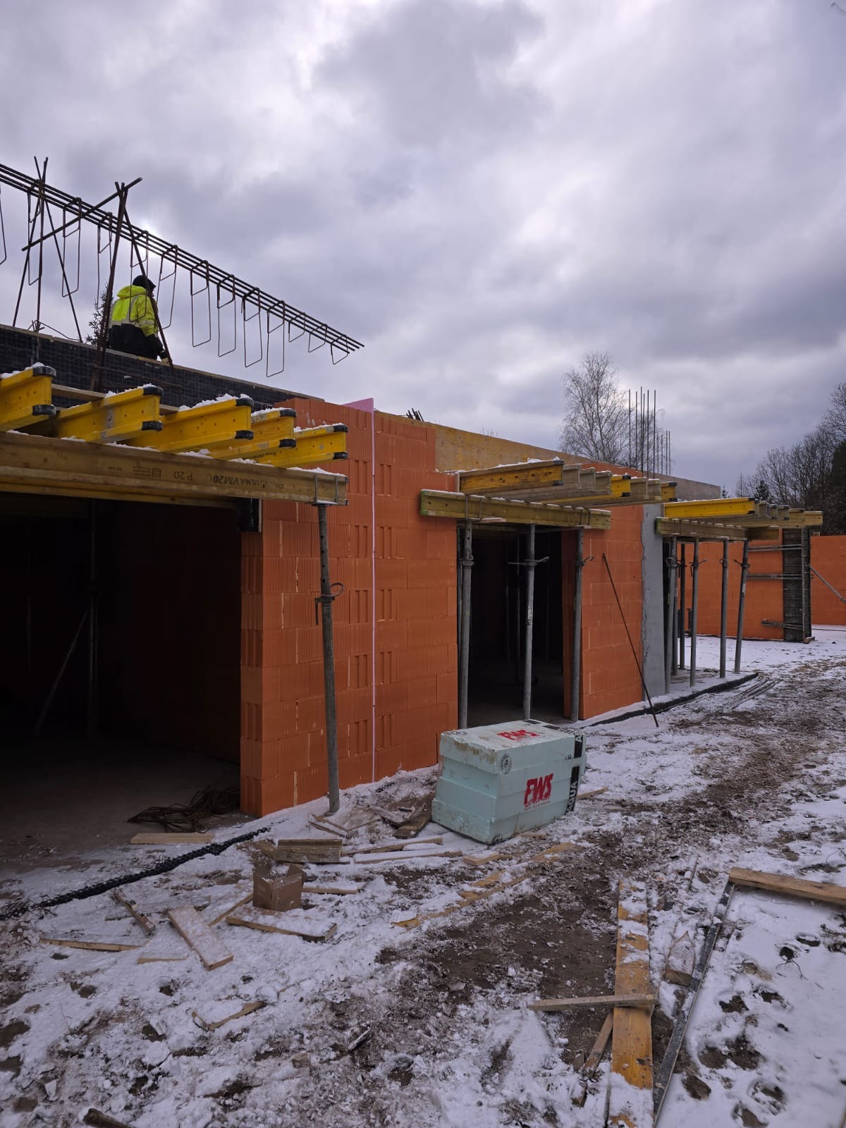 Budowa domu w trakcie zimy: widoczne ściany z pustaków ceramicznych, szalunki pod wylewki stropu, zbrojenie stropu montowane przez pracownika w żółtej kurtce, na ziemi leży śnieg i resztki...