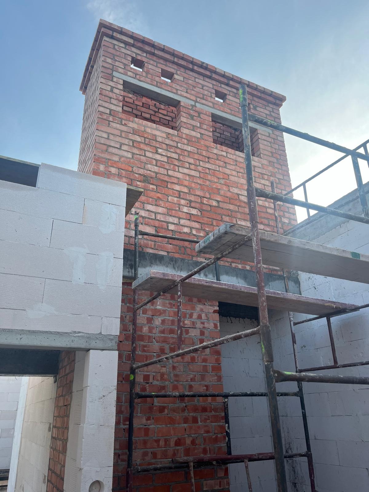 Wznoszenie komina z czerwonej cegły na budowie domu, widoczne rusztowanie i ściany z betonu komórkowego.