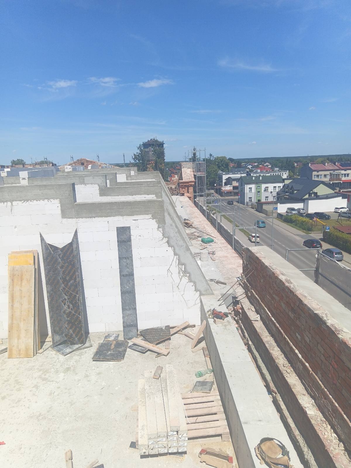 Wysoki kąt widzenia na budowę z białych bloków i czerwonej cegły, widoczne szalunki, narzędzia i panorama miasta w tle pod błękitnym niebem.