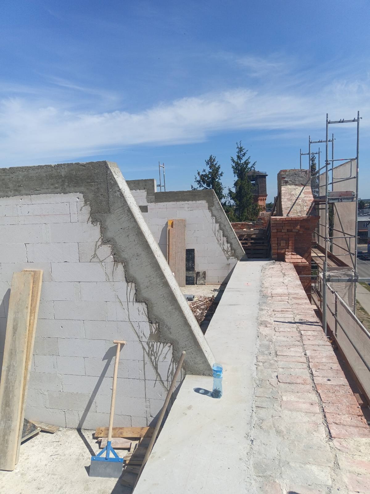 Wznoszenie ścian domu z bloczków, widoczne narzędzia murarskie, fragment rusztowania i butelka wody na murze.