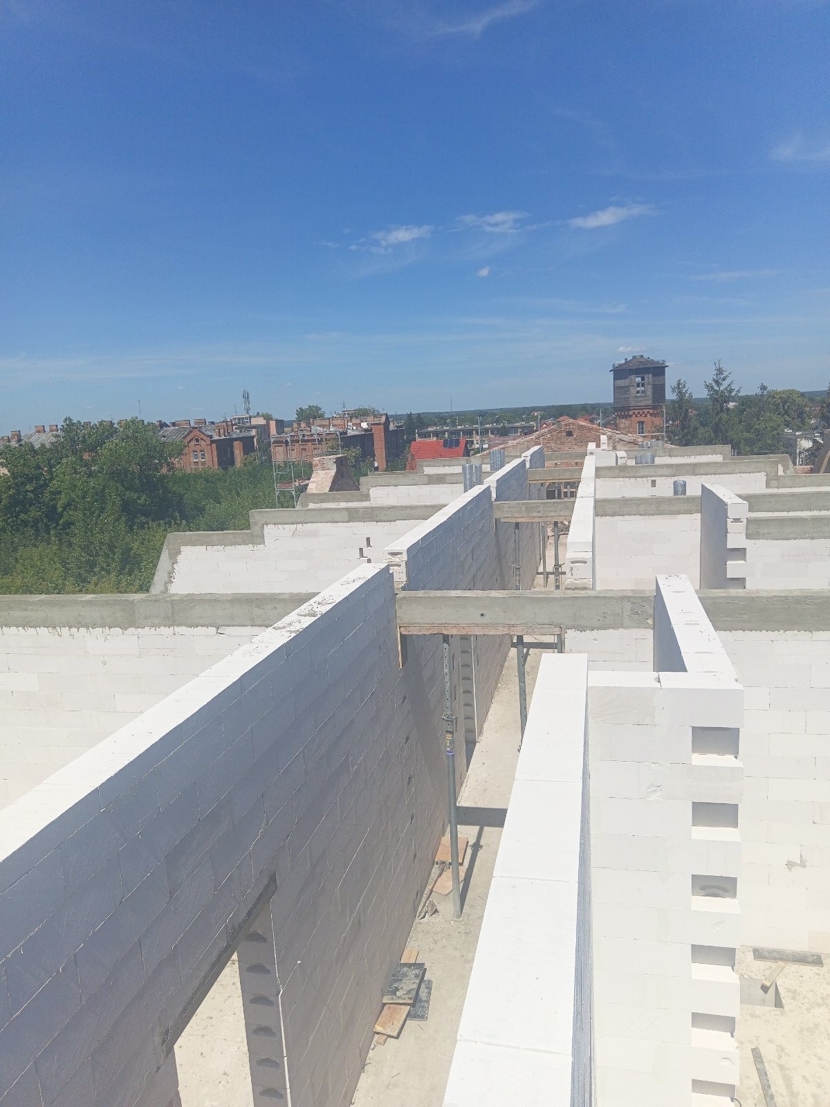 Wznoszenie ścian z białych bloczków na budowie domu, widok z góry, widoczne elementy szalunków i zbrojenia, w tle zabudowa miejska i wieża ciśnień na horyzoncie.