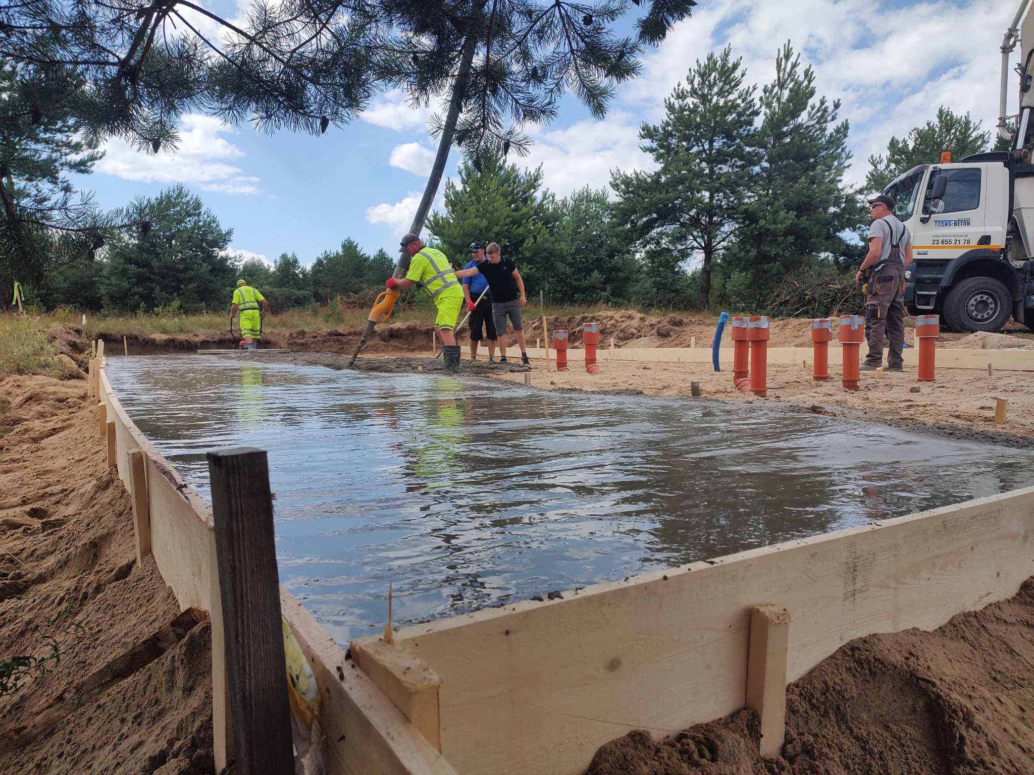 Wylewanie betonu na przygotowany fundament z drewnianym szalunkiem, widoczni pracownicy w jaskrawych ubraniach i betonowóz z wysięgnikiem, teren piaszczysty z drzewami w tle.