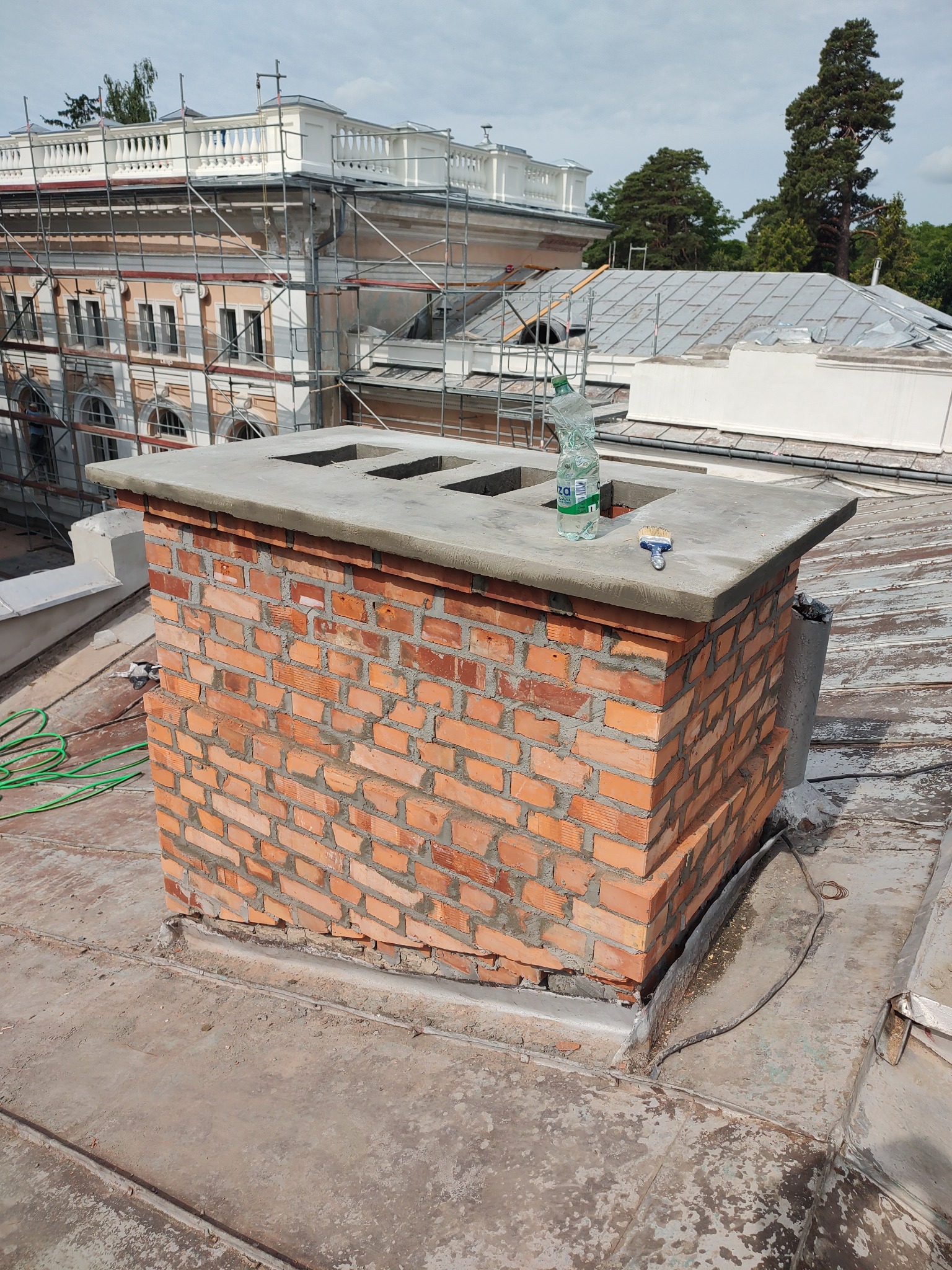 Odnowiony komin z cegły na dachu zabytkowego budynku, z widocznymi rusztowaniami i butelką wody. Detal renowacji komina w zabytkowej architekturze.