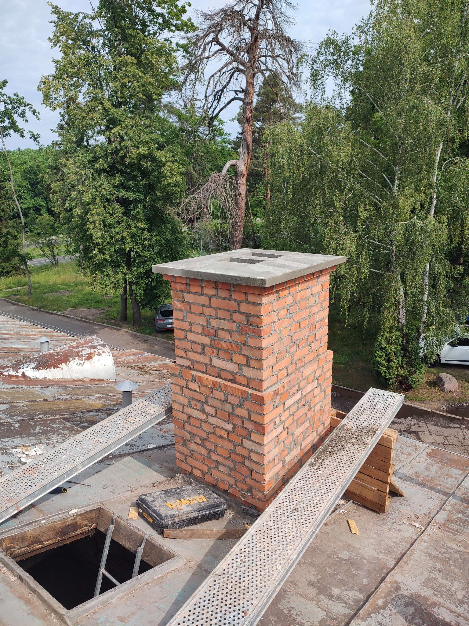 Nowy komin z cegły na dachu budynku w Warszawie, widok z góry. Metalowe pomosty robocze i skrzynka narzędziowa w pobliżu otworu dachowego.