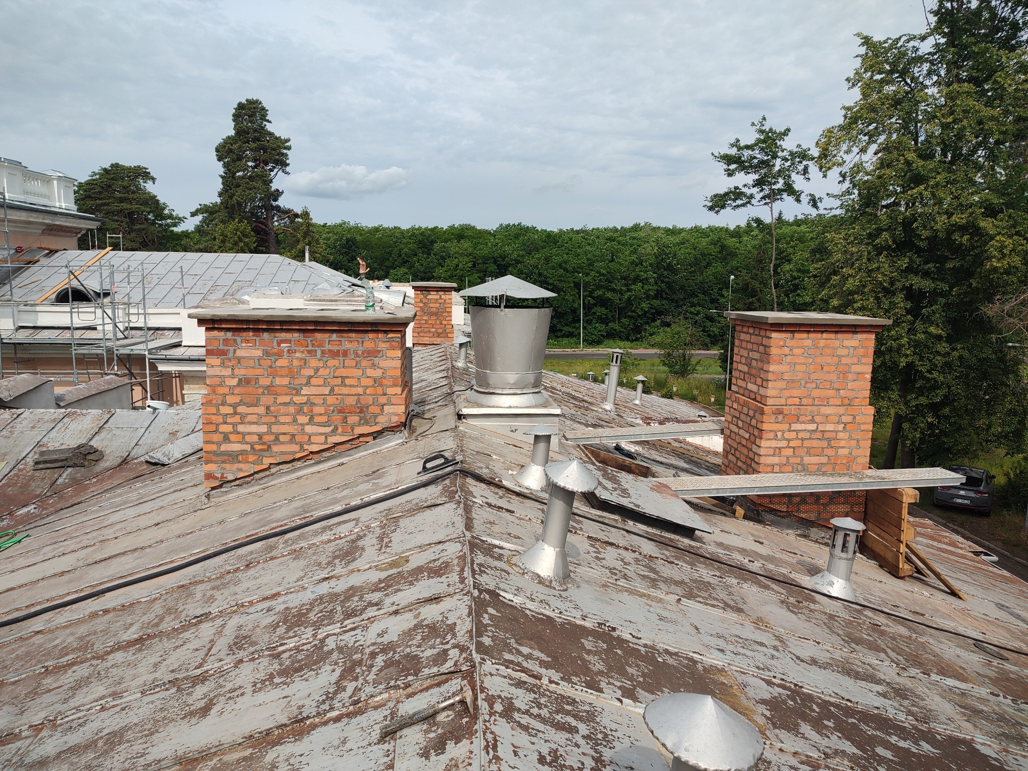Widok z góry na zniszczony dach z ceglanymi kominami i metalowymi elementami wentylacyjnymi, w tle zieleń drzew i fragment budynku w rusztowaniach.