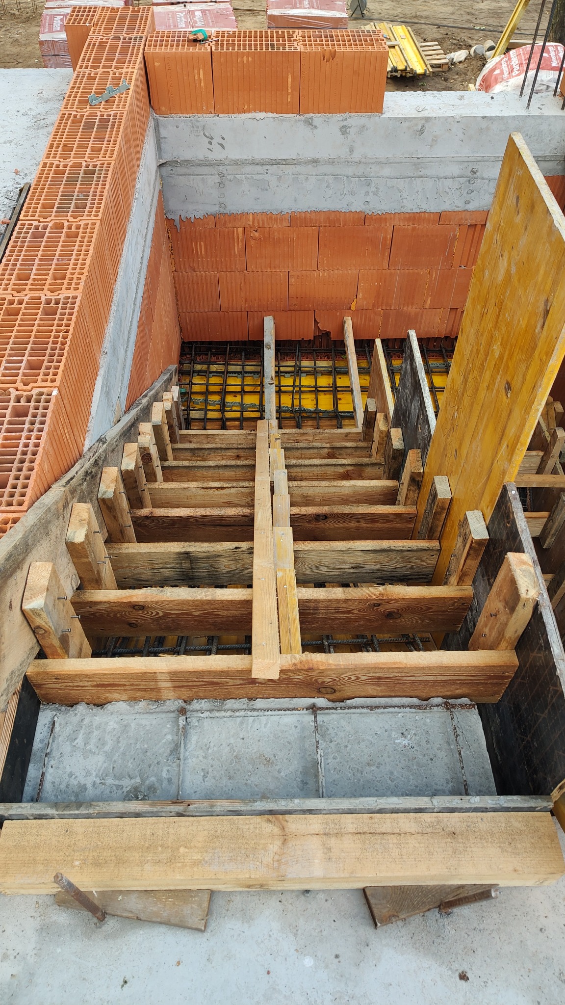 Wykop z deskowaniem schodów, zbrojenie i częściowo wymurowane ściany z cegły dziurawki. Widok z góry na placu budowy w słoneczny dzień.