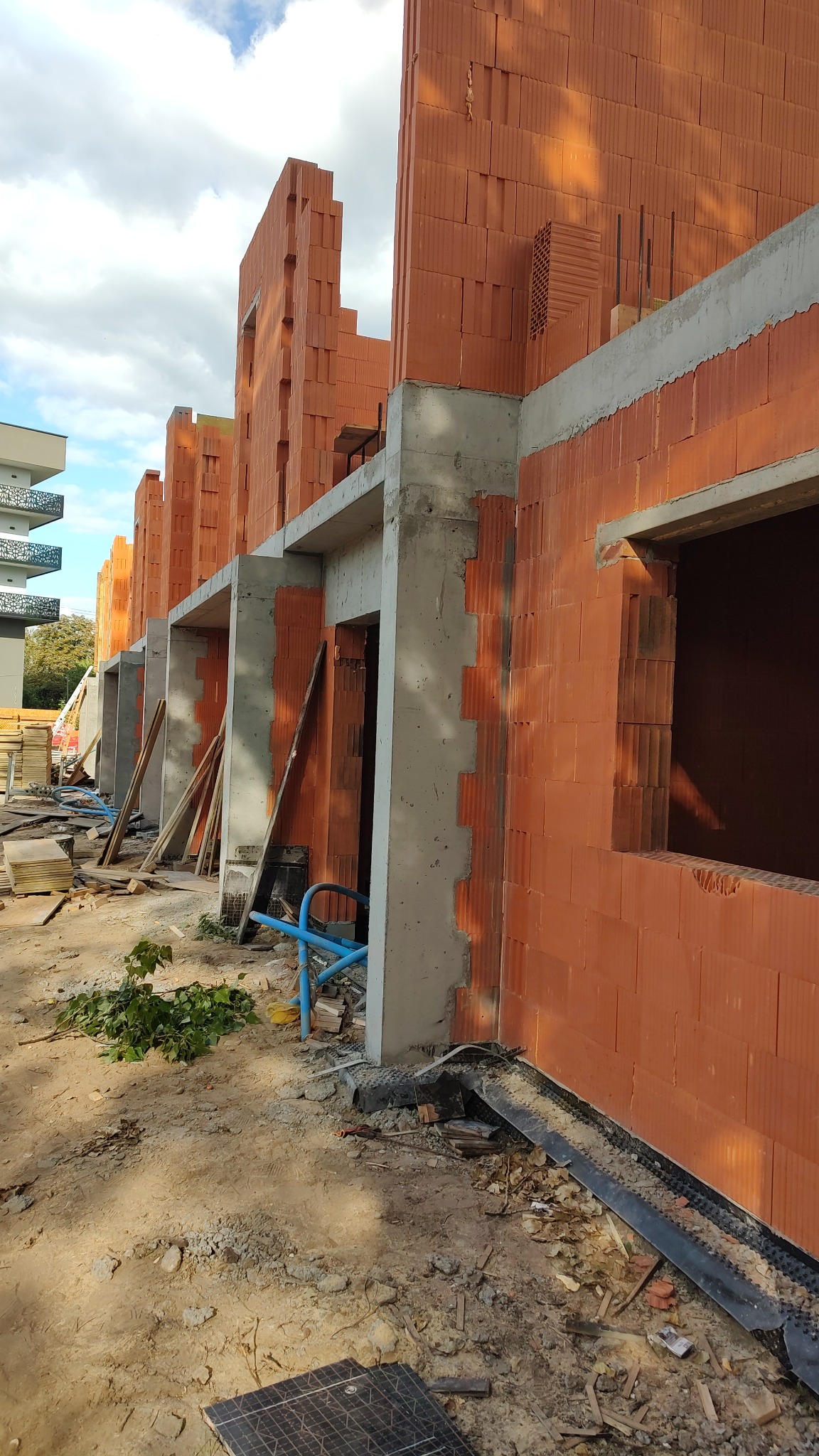 Wznoszenie ścian z czerwonej ceramiki poryzowanej, widoczne betonowe nadproża okien i drzwi. Budowa w toku, na pierwszym planie gruz i fragmenty materiałów budowlanych.