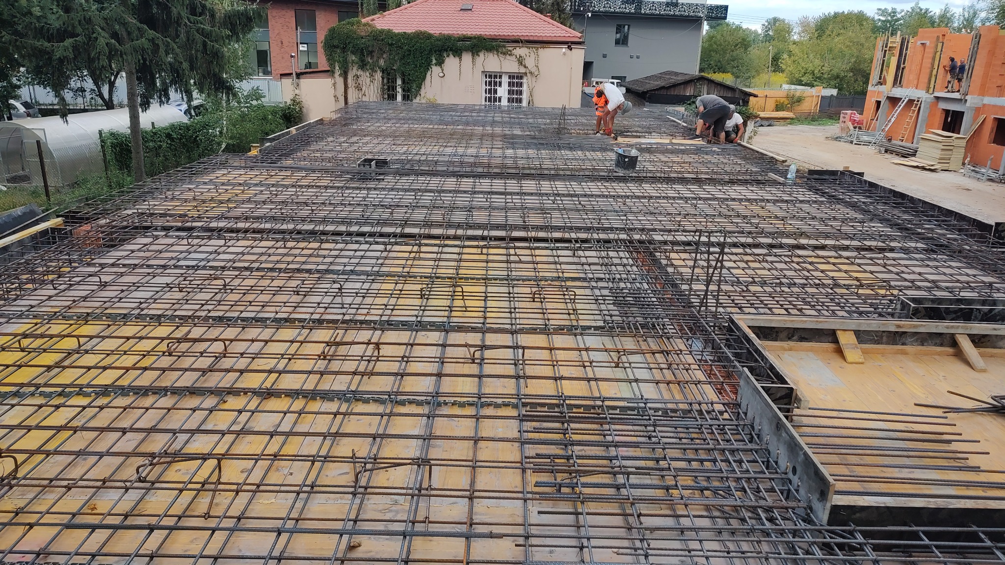 Zbrojenie stropu budynku w trakcie realizacji. Widoczna siatka z prętów stalowych i pracownicy na tle budowy z czerwonej cegły.