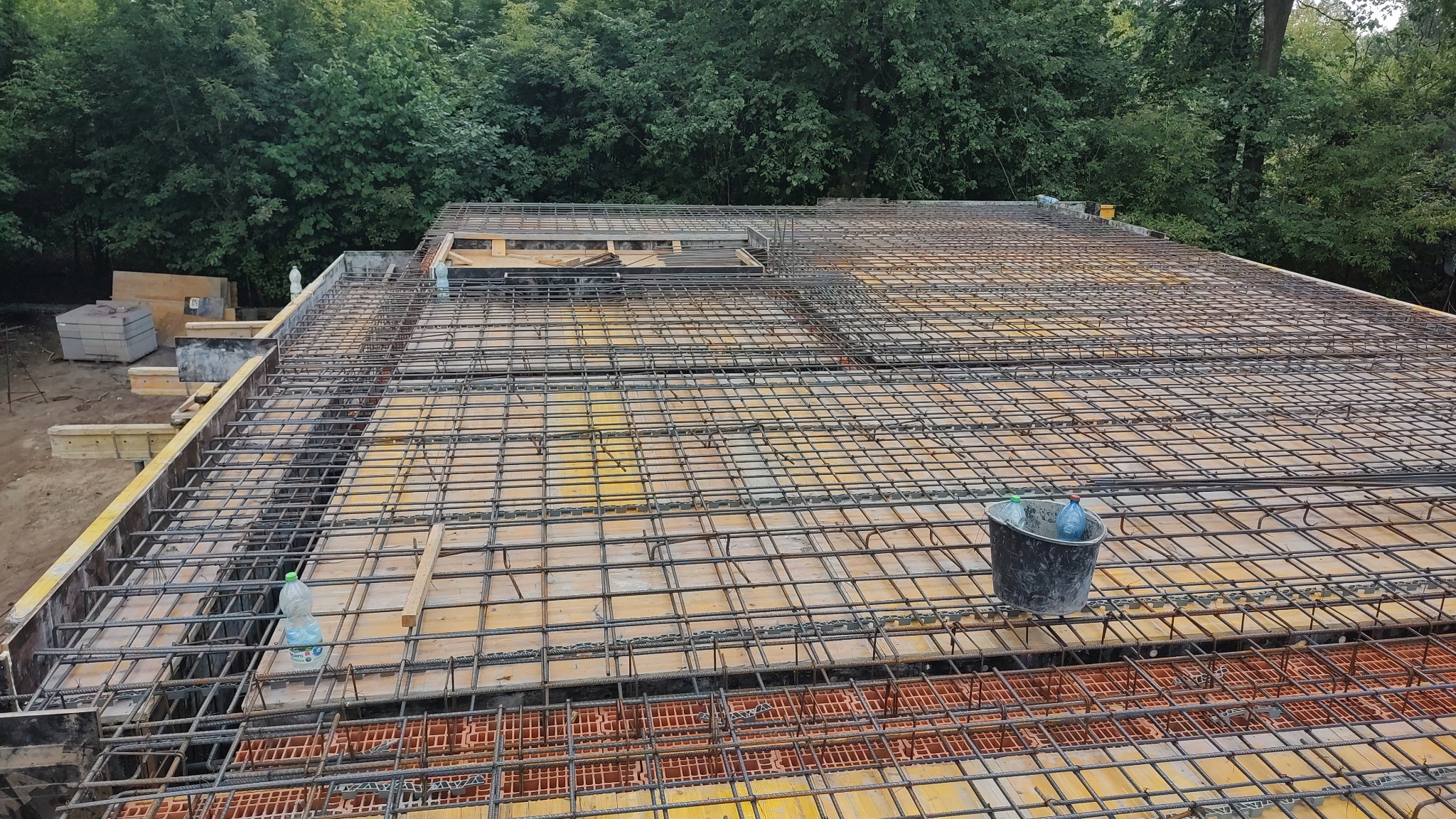 Zbrojenie stropu budynku w trakcie budowy, widoczne szalunki, siatka zbrojeniowa i wiaderko z wodą. W tle zieleń drzew.