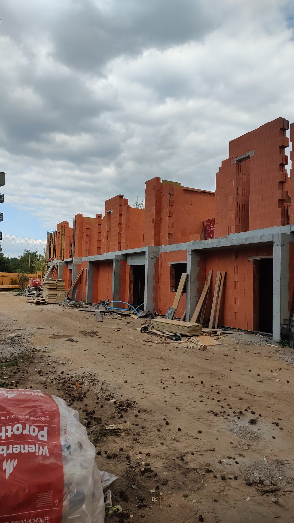 Budowa szeregowców z czerwonej cegły Porotherm. Widoczne pustaki, betonowe nadproża, drewniane deski i drabiny. Szary, pochmurny dzień.