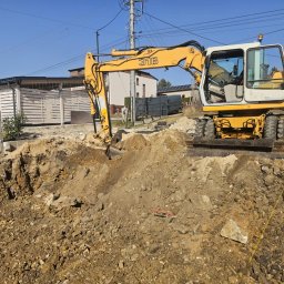 KAS-CAR BARTŁOMIEJ KASPRZAK - Żółto-biała koparka Liebherr 316 przy wykopie w ziemi, widoczne budynki w tle i błękitne niebo.