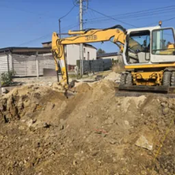 Żółto-biała koparka Liebherr 316 przy wykopie w ziemi, widoczne budynki w tle i błękitne niebo.