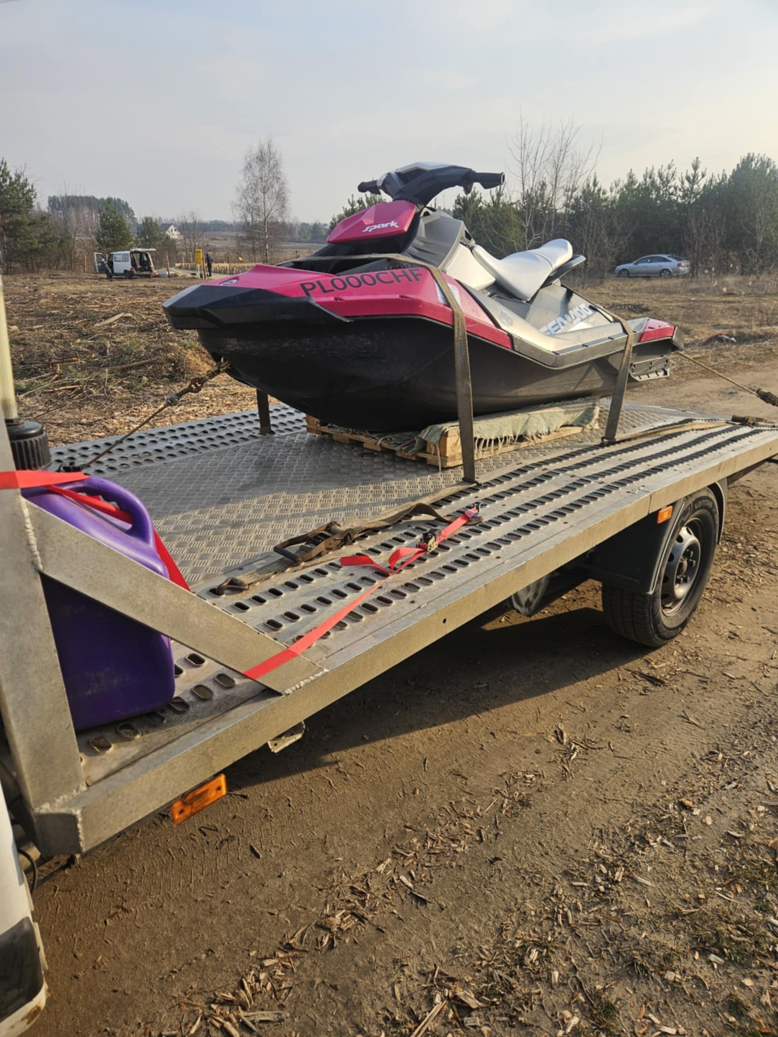 Różowo-biały skuter wodny Sea-Doo na przyczepie lawety zabezpieczony czerwonymi pasami transportowymi, obok fioletowy kanister z paliwem, w tle zalesiony teren i zaparkowane pojazdy.